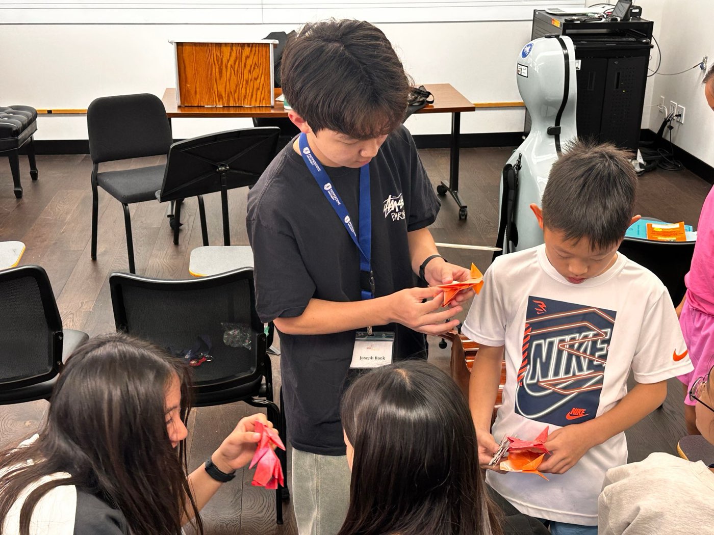 A student volunteer helps a young boy fold orange paper at a community event
